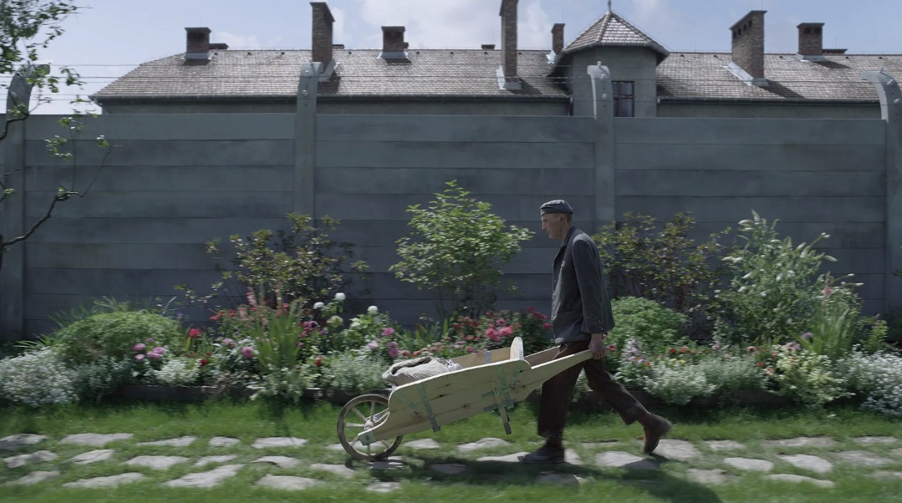 A gardener pushes a wheelbarrow along the garden path with the buildings of Auschwitz beyond the walls.
