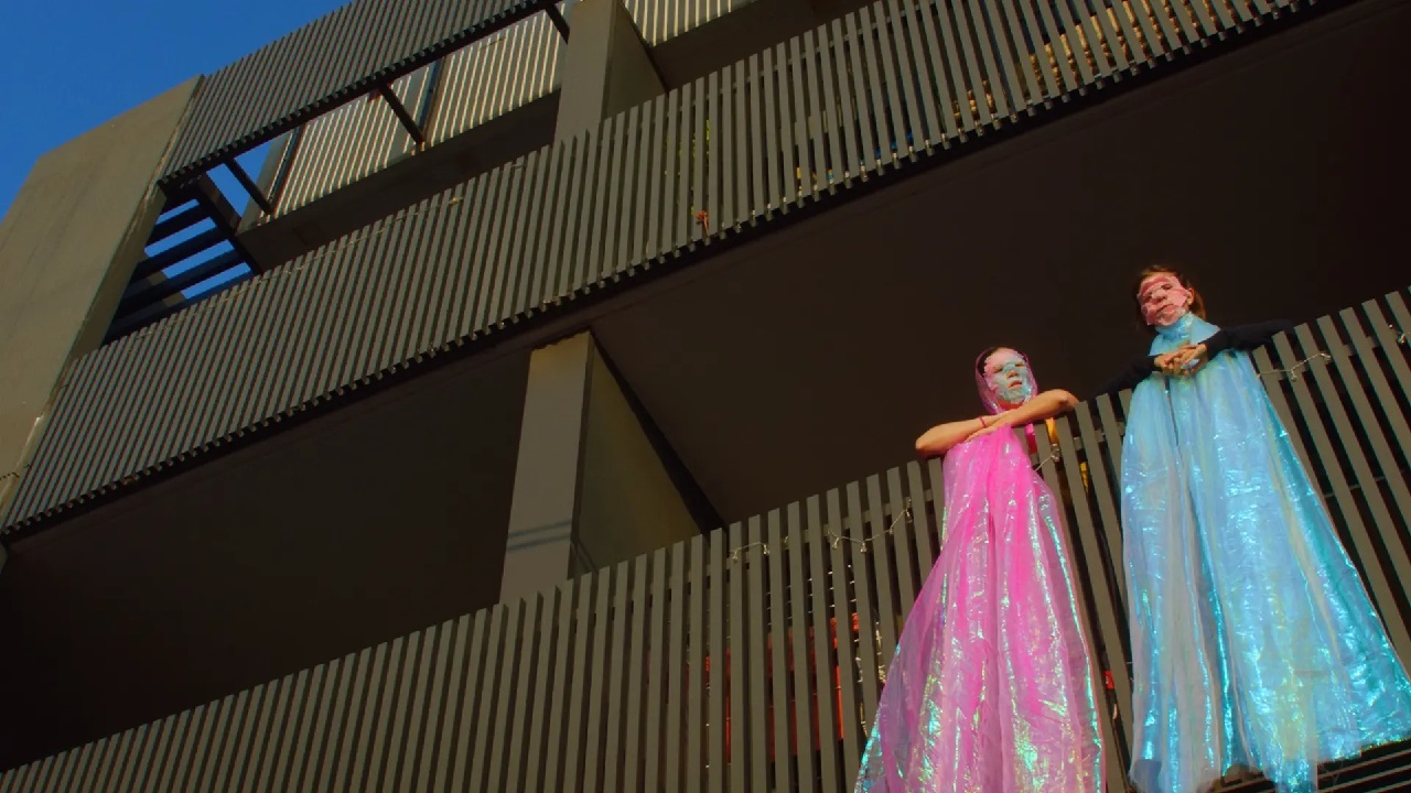 Em (Emmanuelle Mattana) and Jessie (Melissa Gan) stand on Jessie's balcony wearing colourful facemasks and scarves in Fwends.