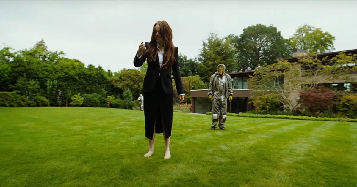 Michelle (Emma Stone) holding a tranquisiler dart shot by Teddy (Jesse Plemons) in a Jennifer Aniston mask in Bugonia.