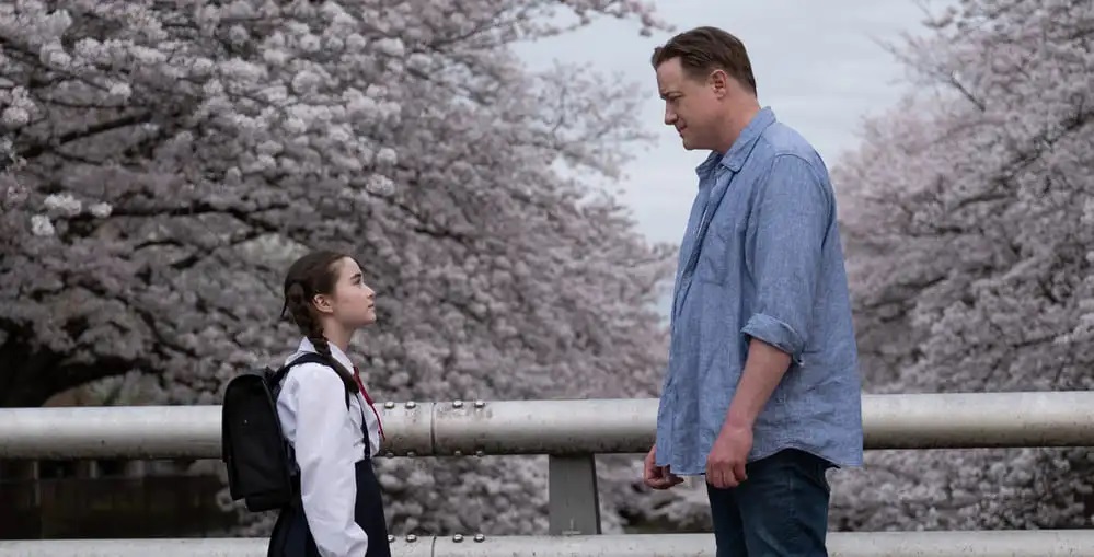 Mia (Shannon Gorman) and her 'father' Phillip (Brendan Fraser) face each other with cherry trees behind in Rental Family.