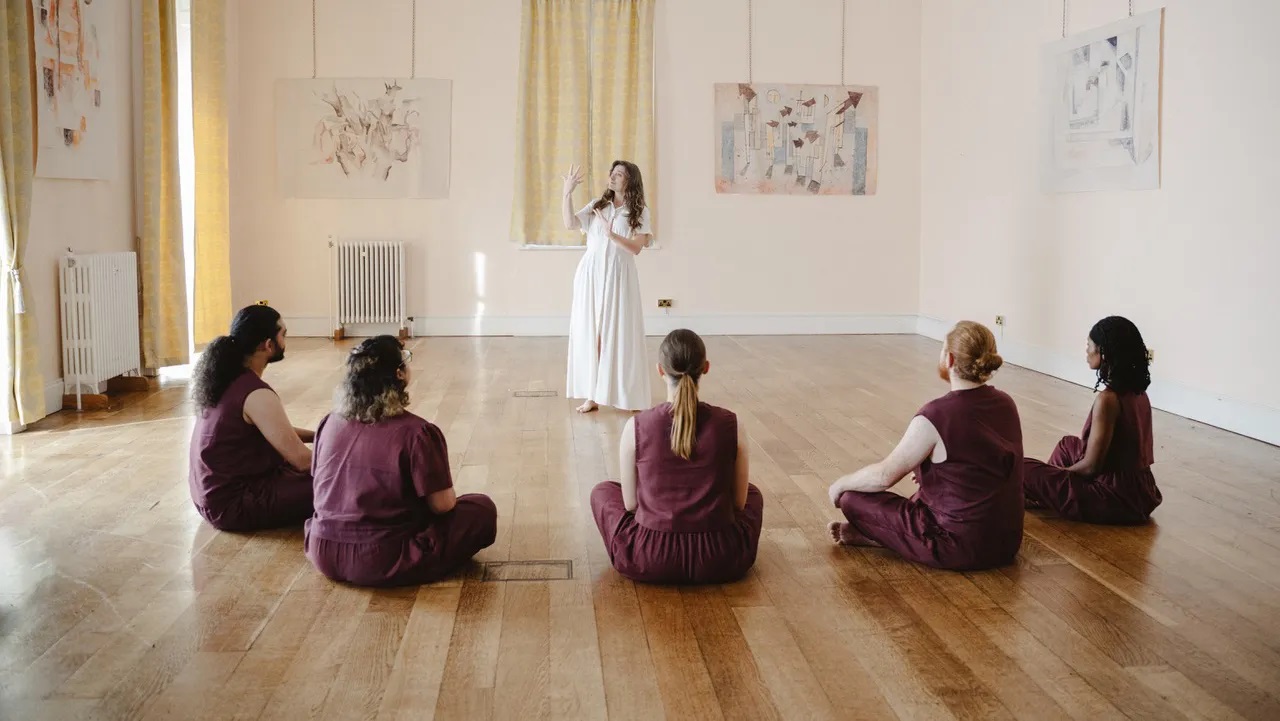 A group of purple clad residents sit on the floor watching a lady in white for the movie Retreat (2025, dir. Ted Evans)